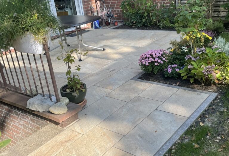 Helle Terrassenfläche mit sandfarbenen Platten in Naturstein-Optik, sauber eingefasst mit dunklem Granit und direkt angrenzenden, bunt bepflanzten Beeten.