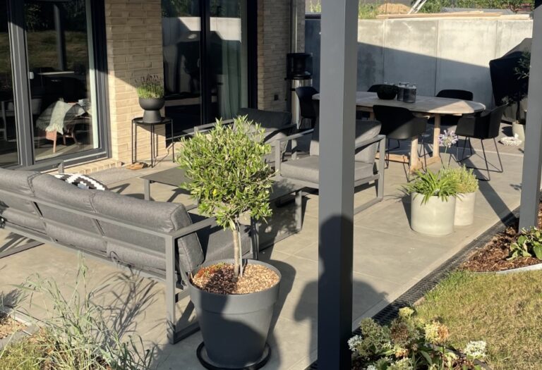 Moderne Terrassengestaltung mit großformatigen, grauen Platten, eingerichtet mit einer gemütlichen Lounge-Ecke und Essbereich, vor einer hellen Klinkerfassade mit bodentiefen Fenstern.