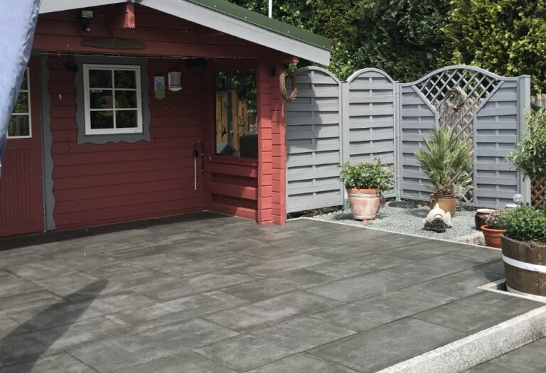 Gepflasterte Terrasse mit dunklen, großformatigen Platten vor einem roten Gartenhaus, seitlich begrenzt durch einen grauen Sichtschutzzaun und dekorative Pflanzkübel.