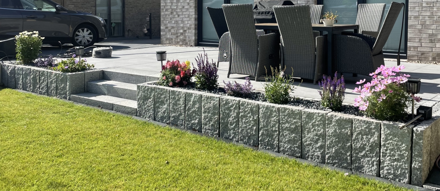 Hochwertige Terrassen- und Beeteinfassung aus massiven Granit-Palisaden, die eine erhöhte Terrasse stützen und gleichzeitig als blühendes Hochbeet dienen.