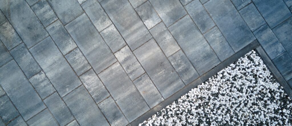 Detailaufnahme moderner Wegegestaltung: Diagonal verlegtes graues Rechteckpflaster, sauber abgegrenzt durch einen Kantenstein von einem Beet mit weißem Zierkies.