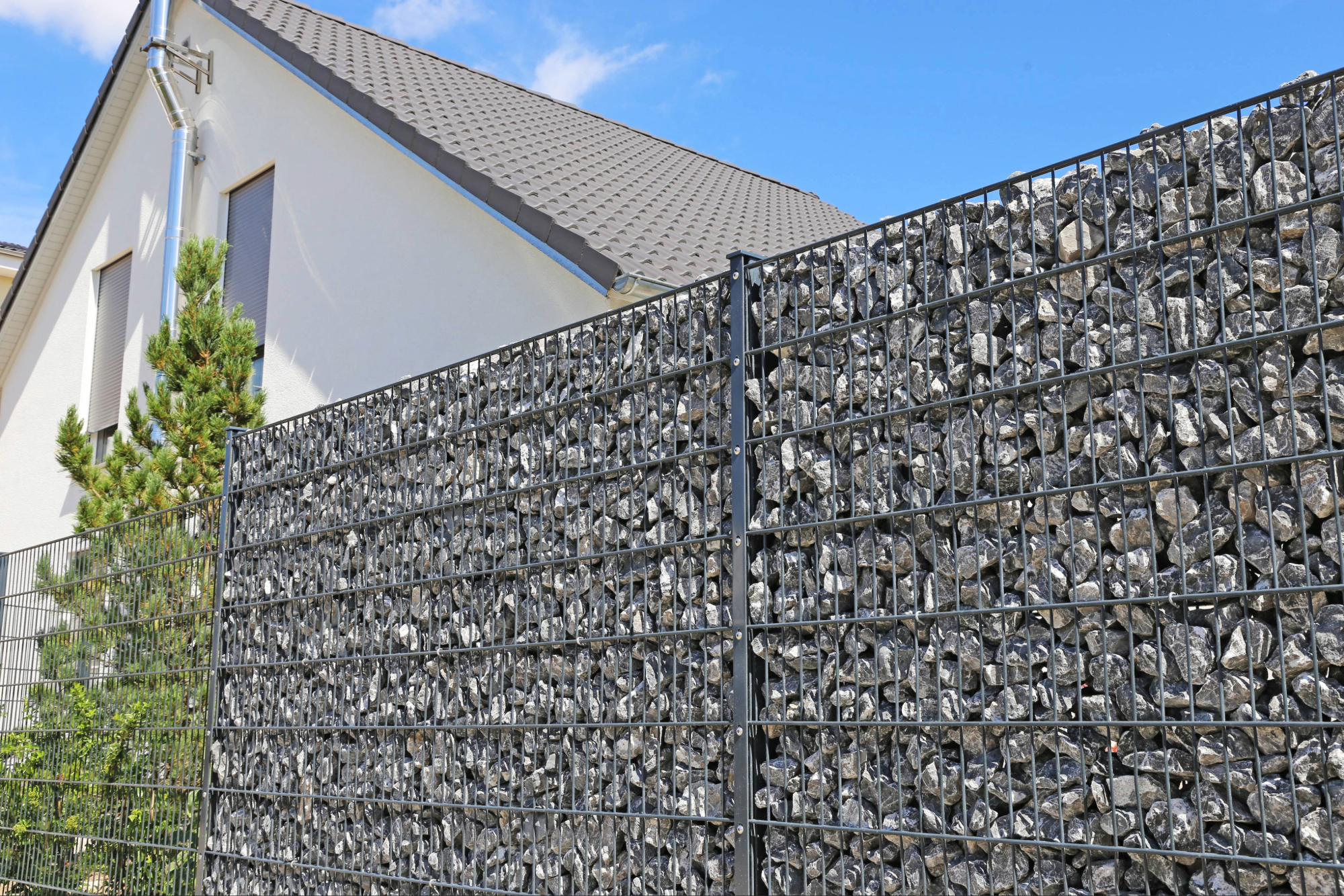 Moderner Gabionenzaun mit Metallrahmen gefüllt mit grauen Steinen neben einem weißen Wohnhaus mit grauem Dach; links eine kleine grüne Pflanze, klarer blauer Himmel.