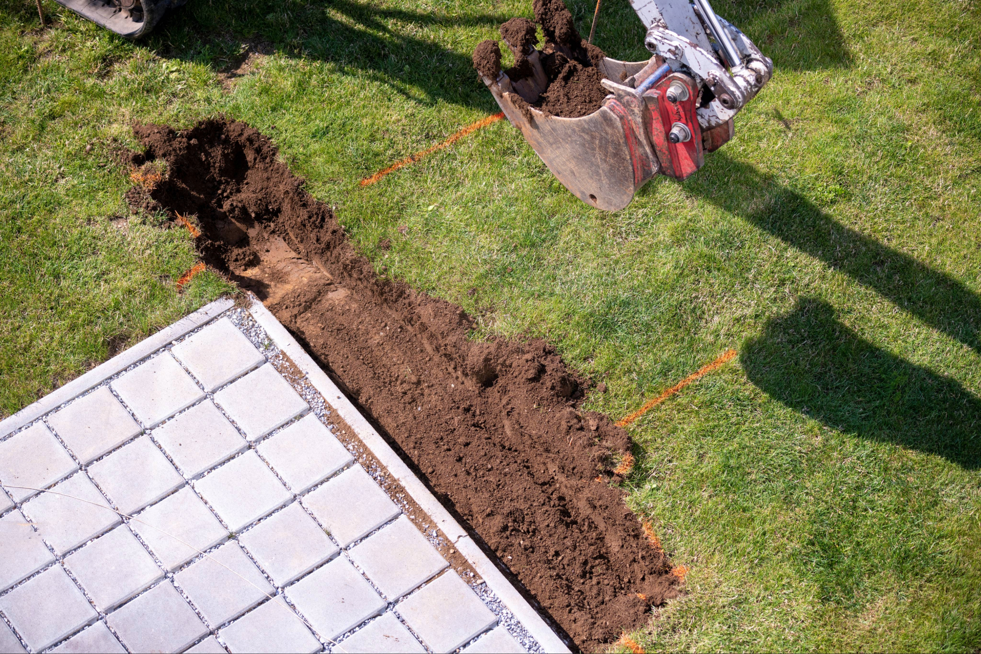 Luftaufnahme einer Baustelle: Bagger schaufelt Erde für Zaunfundament aus; weiße Betonplatten liegen links neben der Ausgrabung, orange Markierungslinie zeigt die Zauntrasse.