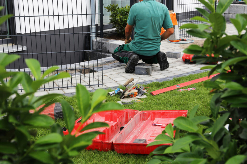 Handwerker in grünem Shirt kauert neben einem schwarzen Metallzaun-Gitterwerk; rote Kunststoffkisten mit Werkzeugen und Material liegen auf dem grünen Rasen, grüne Hecken im Hintergrund.