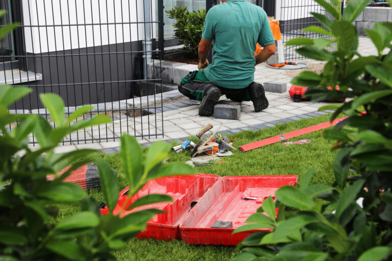 Handwerker in grünem Shirt kauert neben einem schwarzen Metallzaun-Gitterwerk; rote Kunststoffkisten mit Werkzeugen und Material liegen auf dem grünen Rasen, grüne Hecken im Hintergrund.