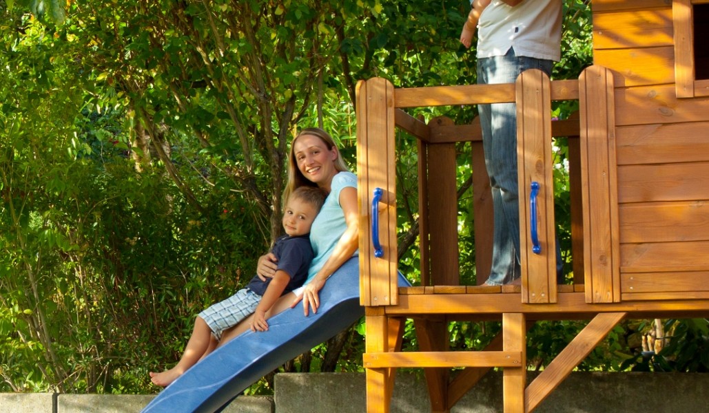 Familienzeit im Garten: Mutter und Sohn rutschen gemeinsam von der blauen Rutsche eines massiven Spielhauses aus Holz, umgeben von grünen Bäumen.
