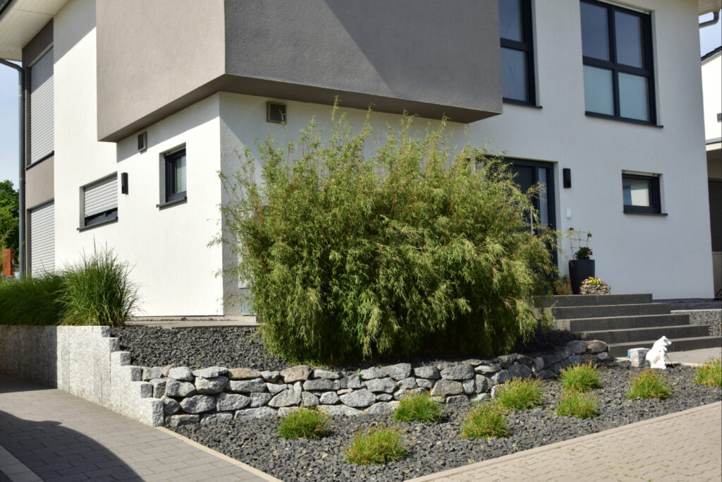Moderner Vorgarten mit Natursteinmauer, Bambus, Ziergräsern und dunklem Ziersplitt vor einem Wohnhaus.